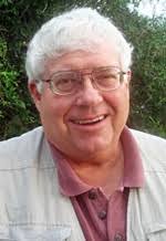 A closeup of a smiling man with gray hair, glasses, brown shirt and a white jacket.