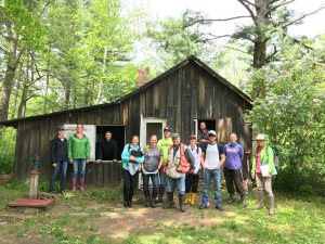 12 people are standing in from of a small, old, wooden structure with a peaked roof on one side. Large trees surround the structure.