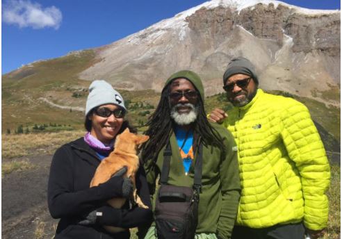 Three Black people wearing sunglasses, jackets, and knit caps are standing facing the camera on a mountain trail. A young woman on the left holds a small dog with golden fur, The man in the center has long dark hair and a gray beard. He is wearing a dark green hooded jacket and carrying a satchel around his neck. The man on the left is the tallest and is wearing a bright yellow down jacket.