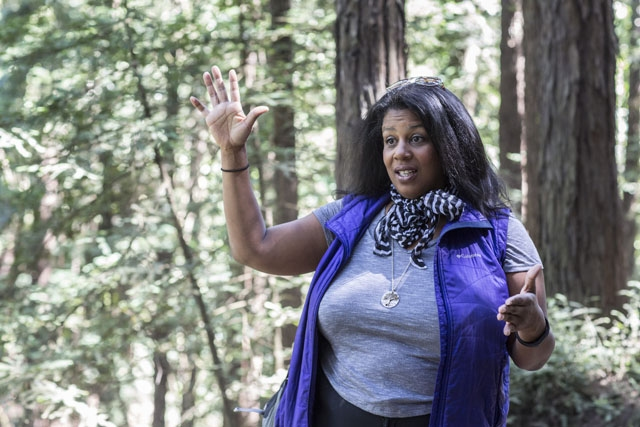 A Black woman wearing a gray t-shirt and a purple vest with a striped scarf around her neck is standing in a forest and gesturing with her hands. Her right arm is raised, bent at a right angle at the elbow with her palm facing forward. Her left arm is at her side, also bent in a right angle at the elbow, with her palm facing left.