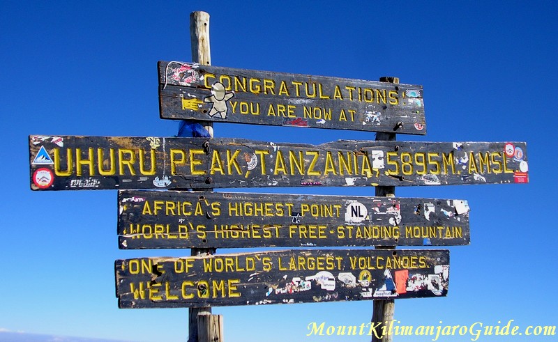 Worn wooden sign with 3 horizontal planks carved with yellow painted words. The top plank reads, "UHURU Peak Tanzania, 5895M. AMSL." The middle one reads, "Africa's Highest Point. World's Highest Free-Standing Mountain." The bottom one reads, "One Of The World's Largest Volcanoes. Welcome." People have placed a variety of stickers on the sign.