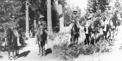 5 black soldiers on horses are lined up side by side on a forest trail and hillside. 