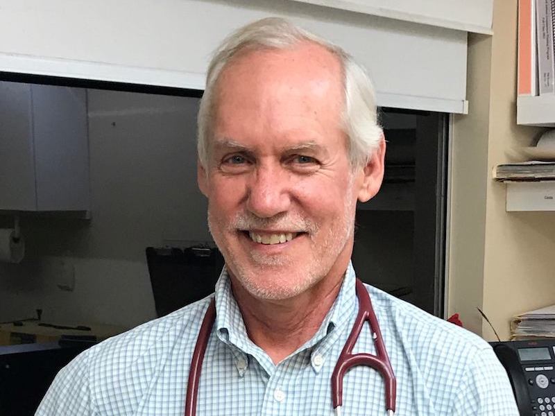 A gray-haired man wearing a blue and white checked shirt, and a stethoscope around his neck, is smiling at the camera.