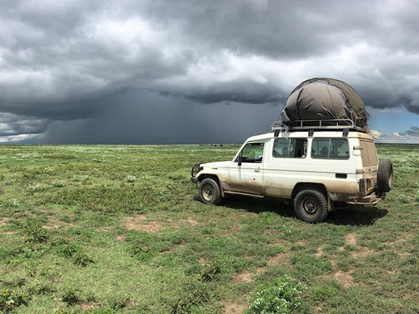 A dirty white Land Rover with large, gray balloon-like object used for wildlife tracking on top, is parked on grassy plain with dark clouds and heavy rain in distance.