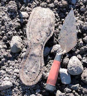 Technology in archaeology in action: old leather boot sole is lying on lumpy dirt, next to a metal trowel with a red handle.