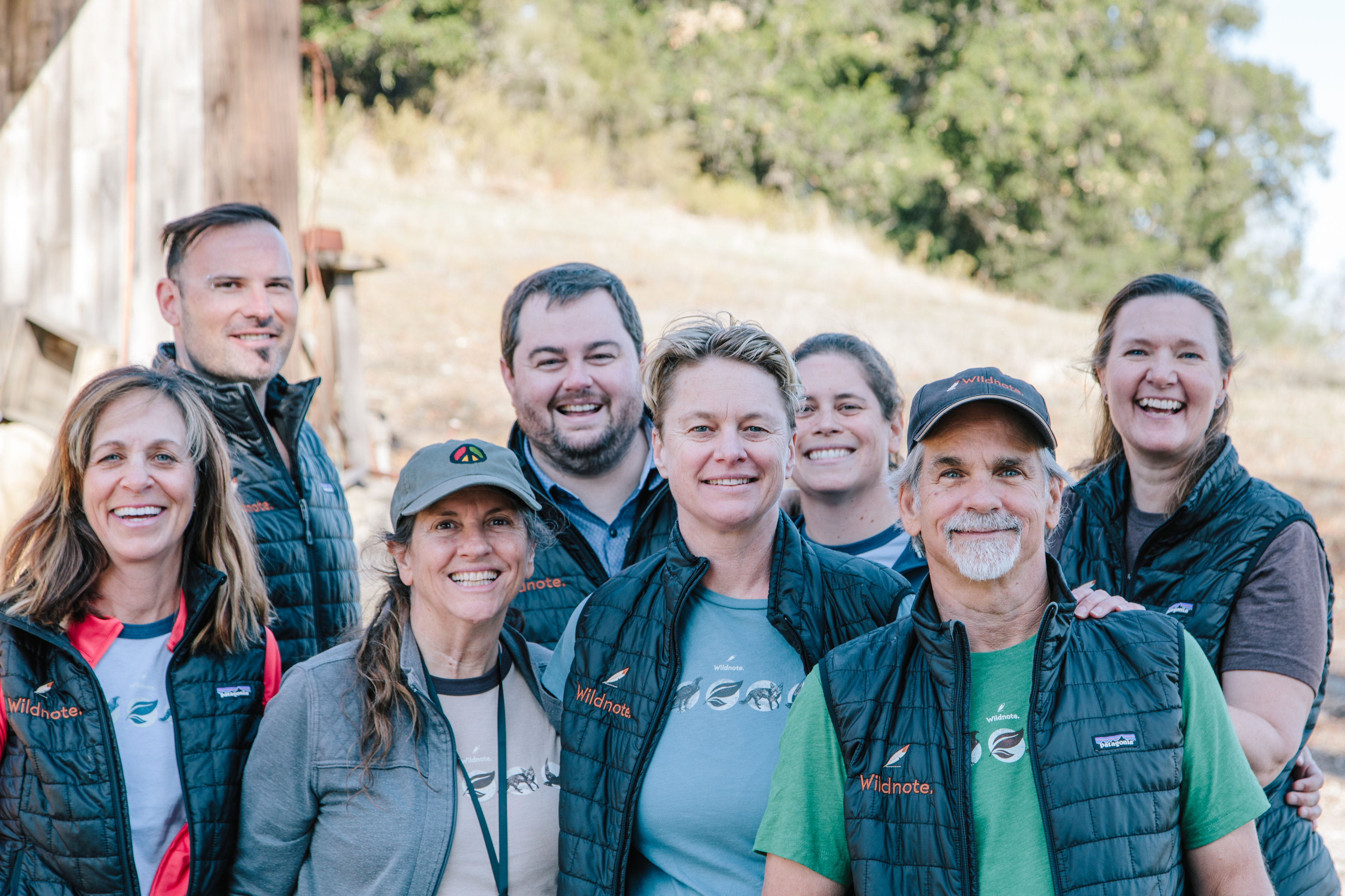 Group of five women and three men smiling and wearing vests bearing the Wildnote name and feather logo