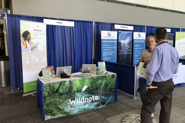Wildnote's field data collection NMEBC demonstration booth draped in rain forest poster, two men conversing in foreground.
