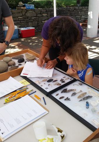 A photo of a woman bending over writing on a piece of paper showing a young girl how to identify and record artifacts next to a case of arrowheads and other cultural resources.