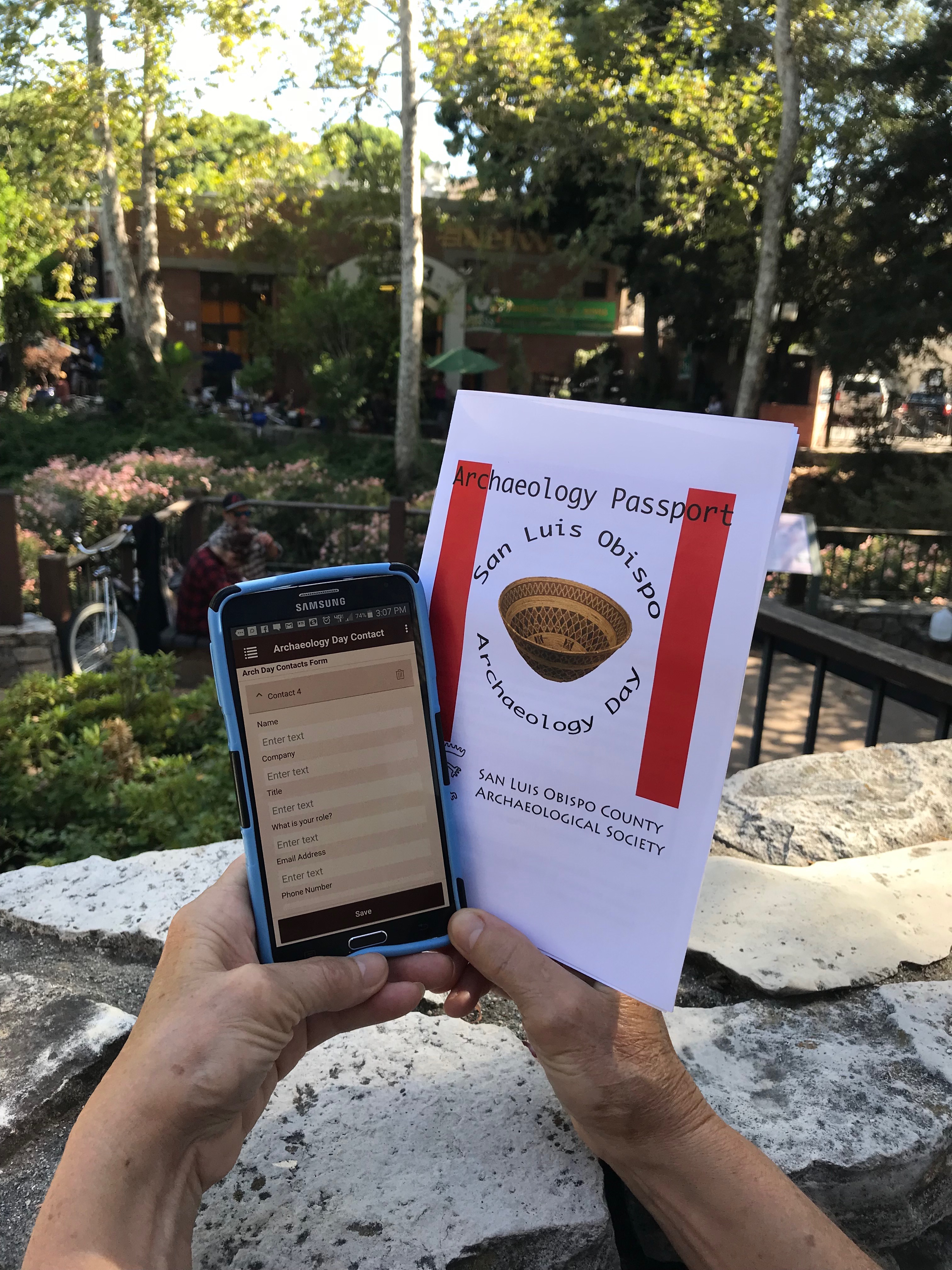 A photo of hands holding a smartphone showing the Wildnote mobile application on it's screen and the cover of an Archaeology Day activity booklet.