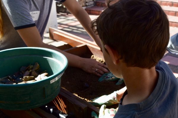 A photo of a female volunteer showing a young boy how to sift soil through a screen to separate out debris that may have archaeological significance.