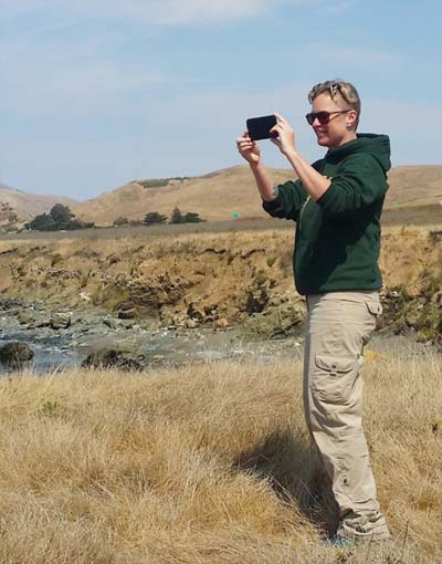 Wildnote CEO Kristen Hazard standing in dry, open country taking photo with mobile phone.