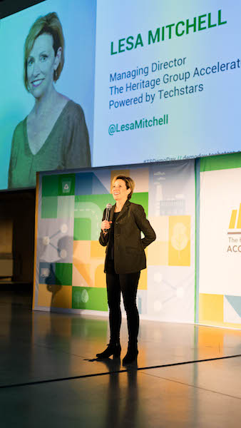 Woman in dark pants and jacket holding a microphone standing on a stage beneath a large screen showing an image of her and her title.