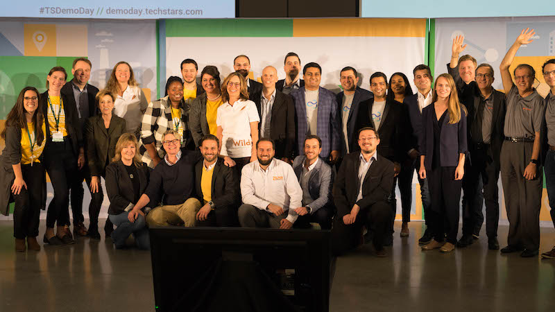 A diverse group of 27 smiling women and men standing and sitting on a stage posing for a group photo